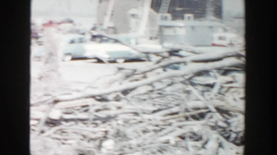 1956 Grand Rapids 8mm frame showing stacked branches or lumber across a cleared site with distant vehicles and buildings.