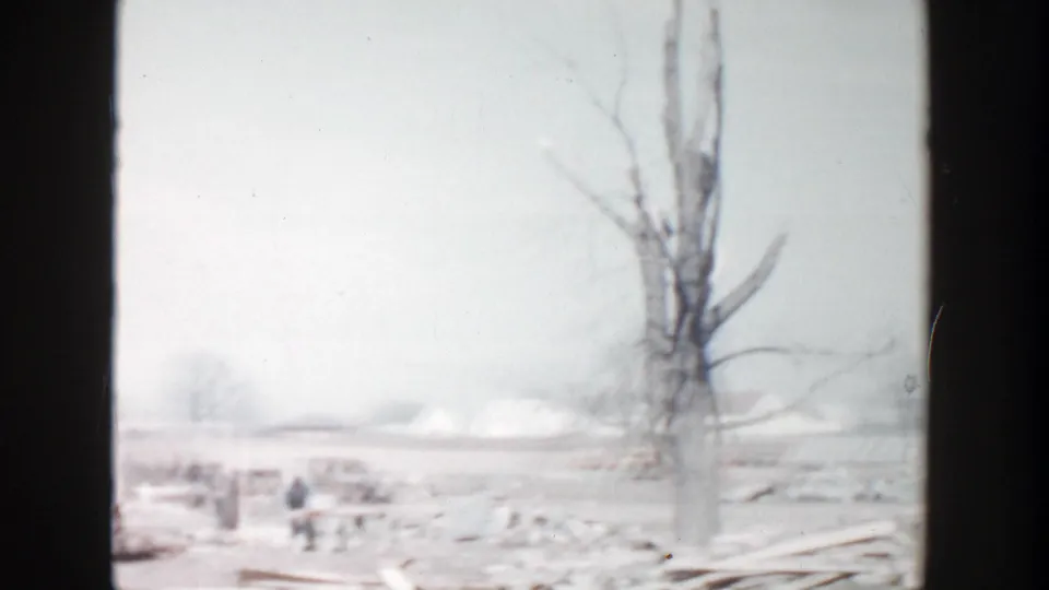1956 Grand Rapids frame with a leafless tree, scattered wood debris, and a small distant person on open land.