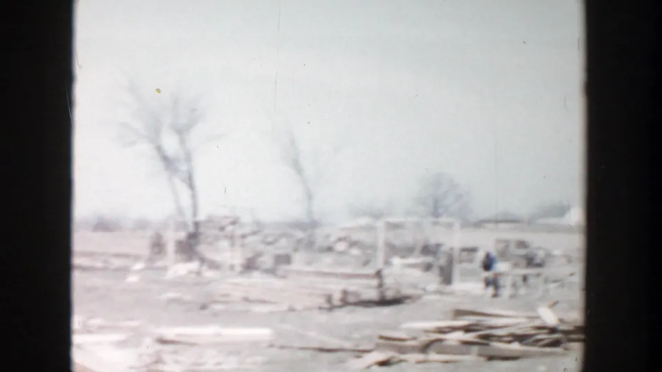 1956 Grand Rapids 8mm frame showing distant workers, parked vehicles, bare trees, and lumber scattered across a cleared site.