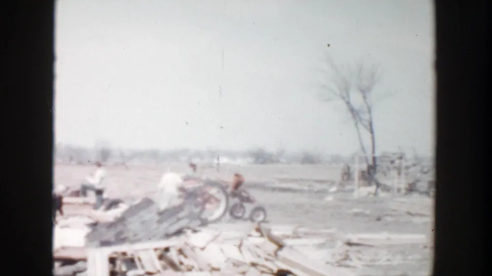 1956 Grand Rapids frame with several distant people near stacked lumber, debris, bare trees, and open ground.