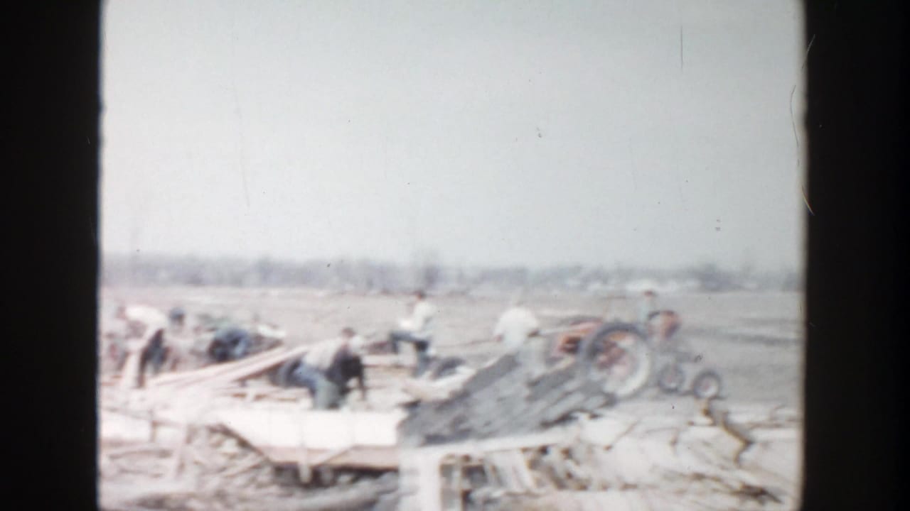 1956 Grand Rapids-area 8mm frame showing workers amid loose boards and debris in a rural field.
