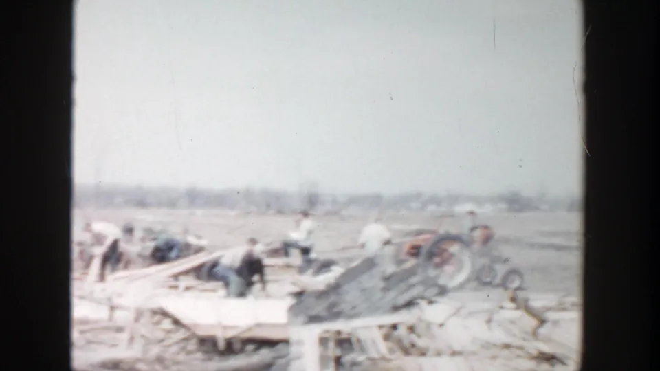 1956 Grand Rapids-area 8mm frame showing workers amid loose boards and debris in a rural field.