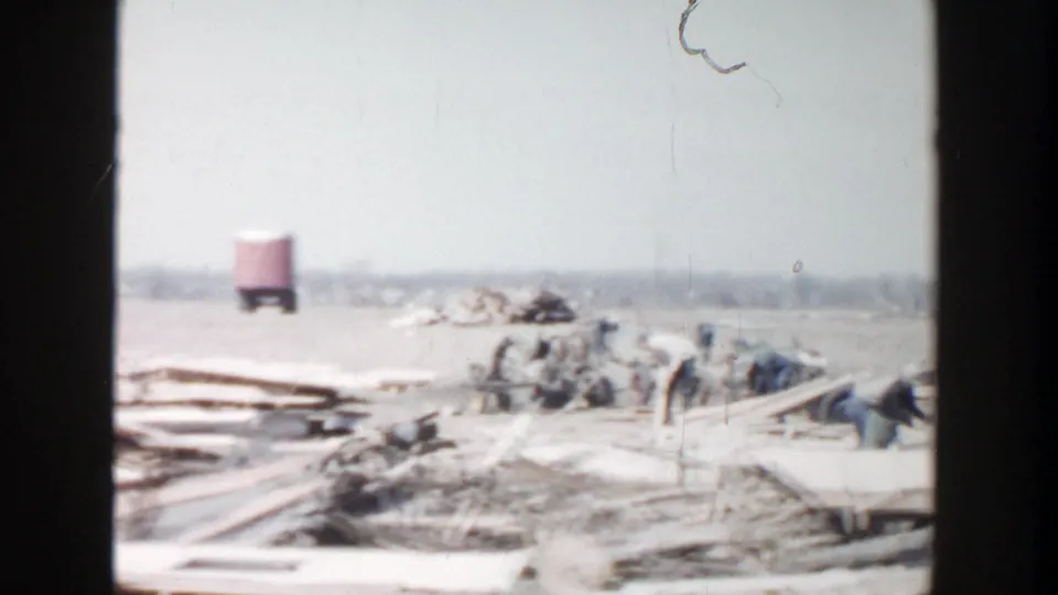 1956 Grand Rapids-area 8mm frame with scattered planks, distant workers, and a red water tower across an open field.