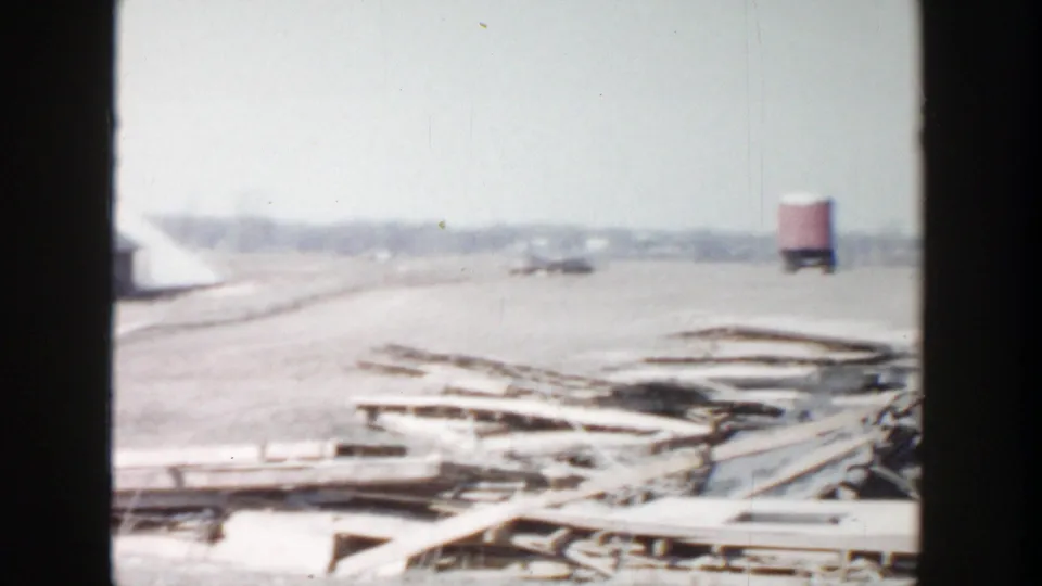 1956 Grand Rapids-area 8mm frame showing foreground boards, cleared ground, and a distant red water tower.