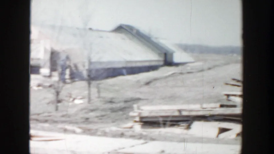 1956 Grand Rapids-area 8mm frame showing a low building, open ground, and stacked boards near the worksite.