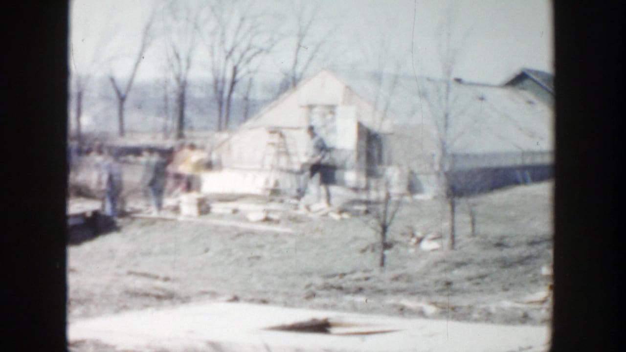 Grand Rapids home-movie frame showing a residential yard, small outbuilding, bare trees, and building materials in 1956 footage.