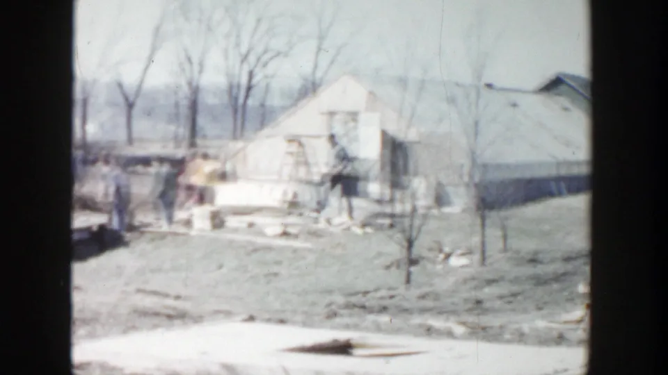 Grand Rapids home-movie frame showing a residential yard, small outbuilding, bare trees, and building materials in 1956 footage.
