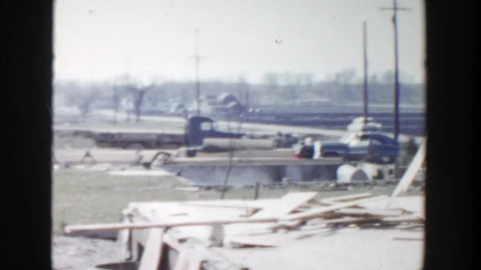 Grand Rapids 1956 aftermath view with scattered boards, utility poles, parked cars, and open ground beyond the damaged structure.