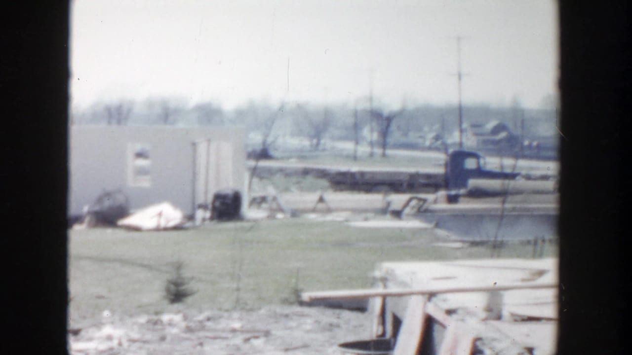 1956 Grand Rapids home-movie frame showing an open work area, low structures, scattered debris, and a distant roadway.