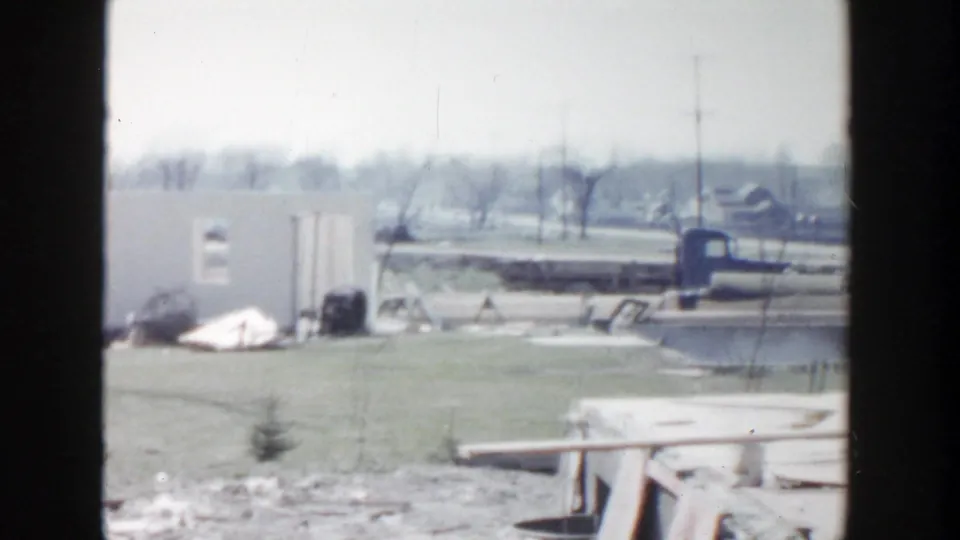 1956 Grand Rapids home-movie frame showing an open work area, low structures, scattered debris, and a distant roadway.