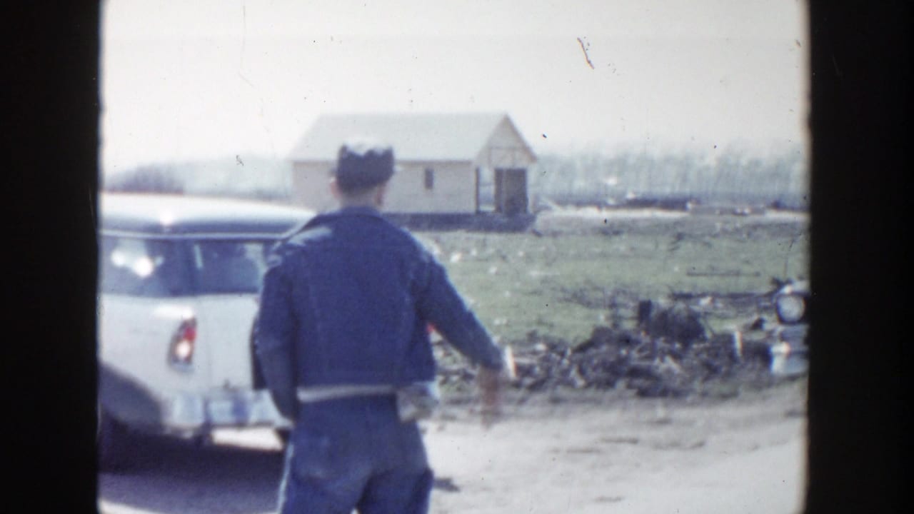 Man in dark jacket stands beside a white station wagon on a rural road.