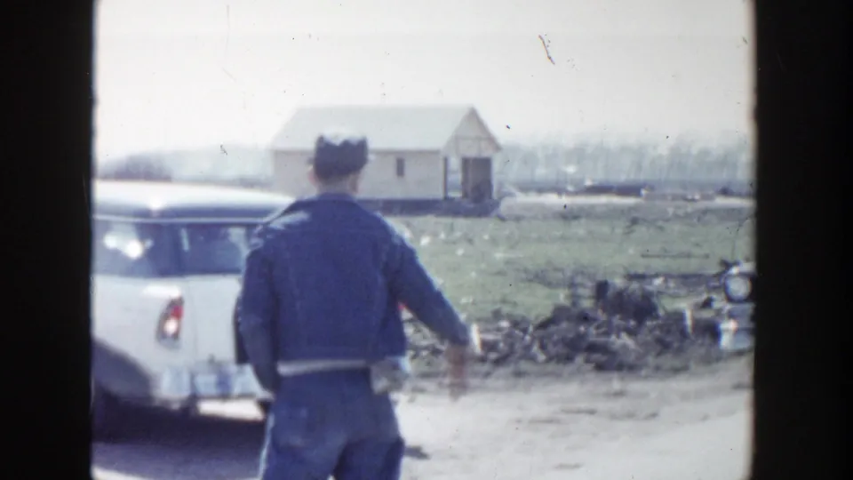 1956 Grand Rapids home-movie frame showing a man in a dark cap and jacket beside a white station wagon near a small rural building.