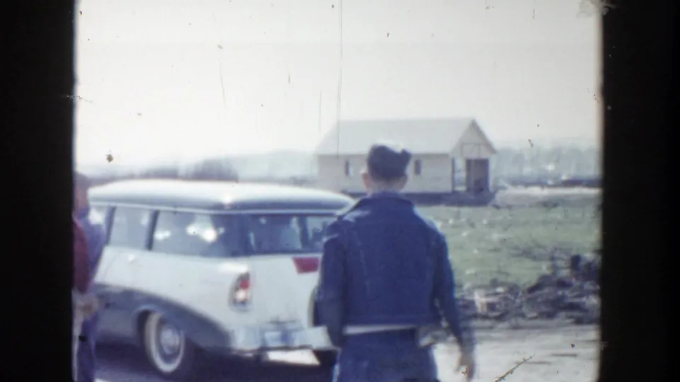 1956 Grand Rapids frame with a man seen from behind near a white station wagon, open roadside ground, and a small building in the distance.