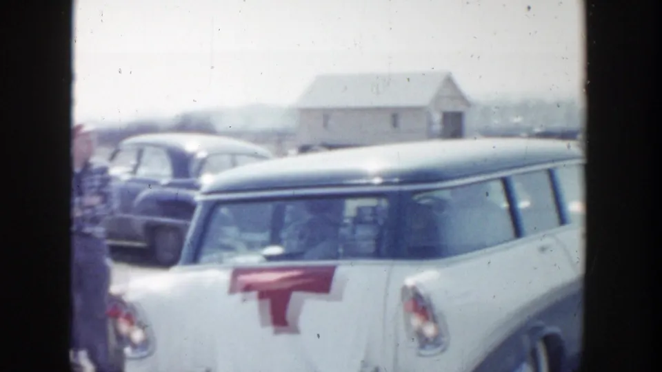 1956 Grand Rapids frame showing a white station wagon with a red rear marking, nearby classic cars, and a small rural building beyond the road.