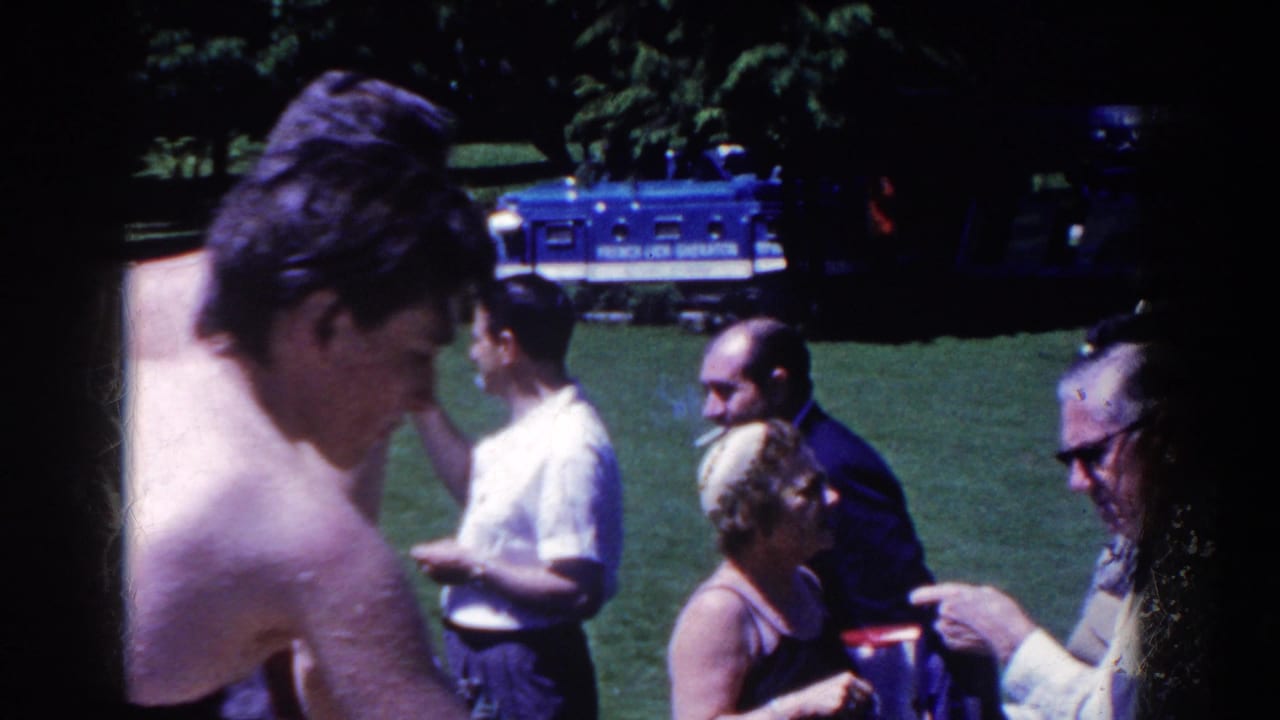 A shirtless man passes in front of adults gathered on grass near a blue bus in the background.