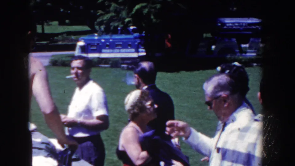 Adults in summer clothing stand on a bright lawn in French Lick, Indiana, with cigarettes, cups, and a blue bus visible.