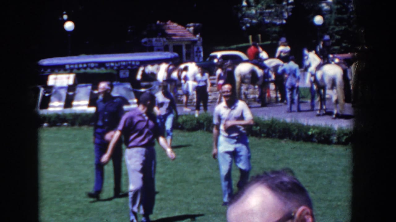 1958 French Lick frame showing adults crossing a lawn while horses, handlers, a carriage area, and a blue tour vehicle appear behind them.