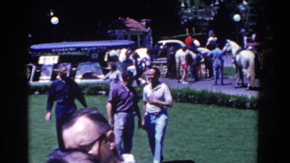 French Lick, Indiana home-movie frame from 1958 showing two men crossing a resort lawn as horses and a blue tour vehicle remain in the background.