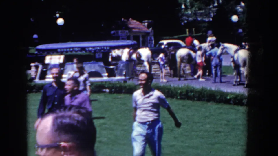 1958 French Lick frame showing adults walking past the camera on grass with horses, riders, and a tour vehicle in the background.