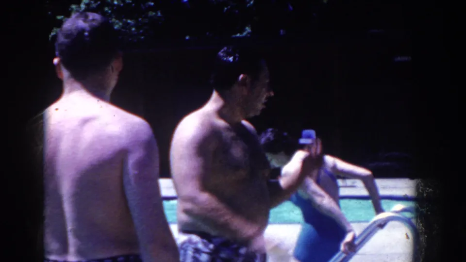Men in swim trunks gather beside the public pool in French Lick, Indiana, with a swimmer and pool railing visible nearby.