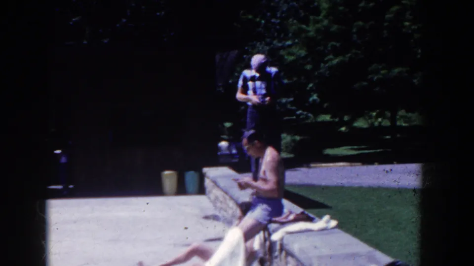 1958 poolside home-movie frame with adults by a low stone wall, shaded park trees, and bright concrete paths.
