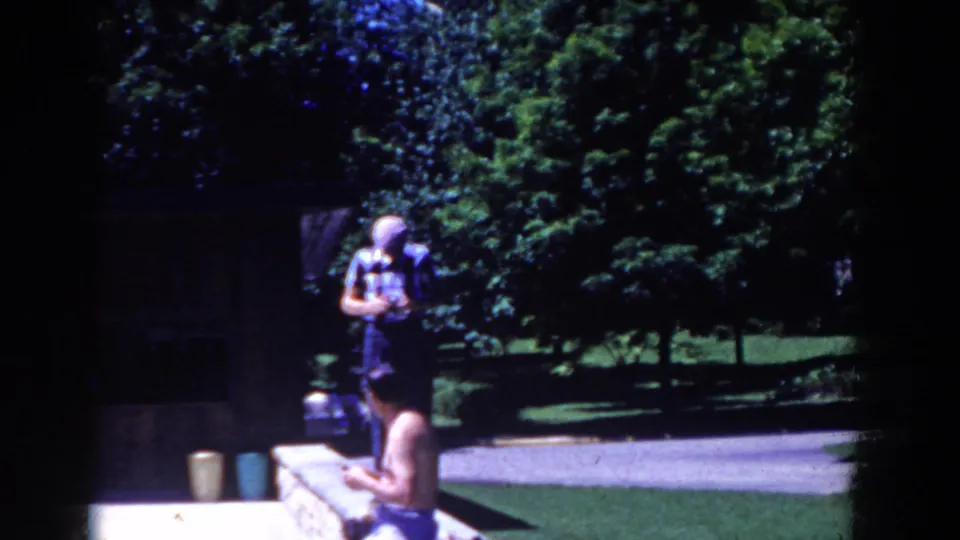French Lick 1958 frame showing a seated poolside adult, another standing near the path, and dense green trees beyond.
