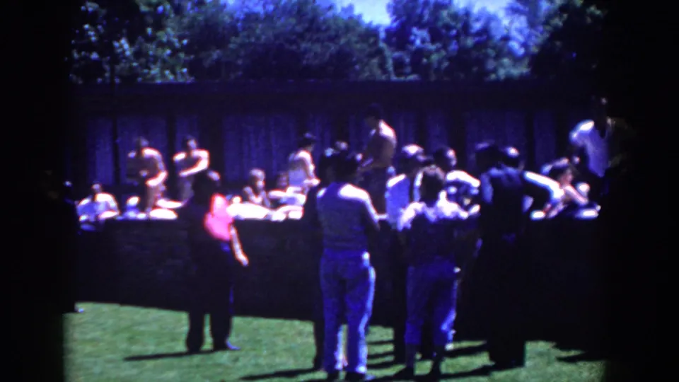 Spectators and participants cluster around a low wall during an outdoor catch activity in the 1958 French Lick footage.