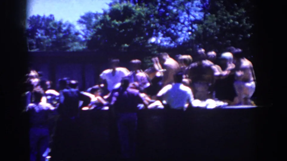 1958 French Lick home-movie frame showing men socializing around a long table beneath trees in bright summer light.