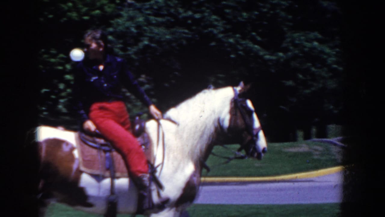 A rider in red pants sits on a white and brown pinto horse in French Lick, Indiana, with park greenery and road behind.