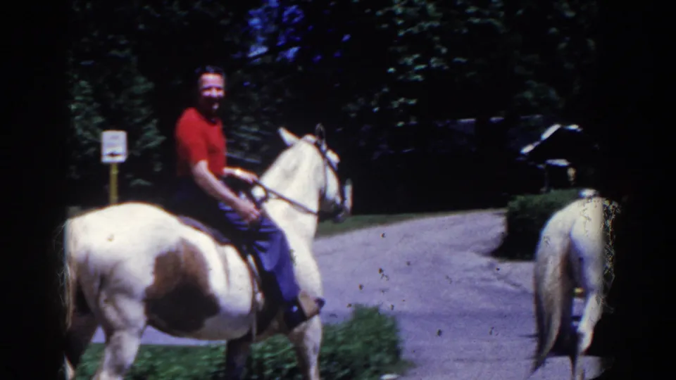 1958 French Lick home-movie frame showing an adult rider in a red shirt on a white-and-brown horse near trees and a path.