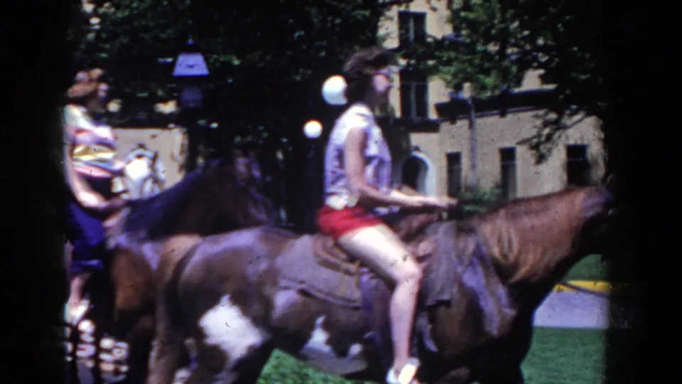 1958 French Lick frame showing women on dark horses, with lawn, lamp posts, trees, and a building in the background.