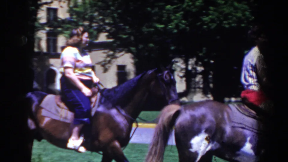 1958 French Lick home-movie frame with riders on dark horses crossing the foreground near trees, lawn, and a building.