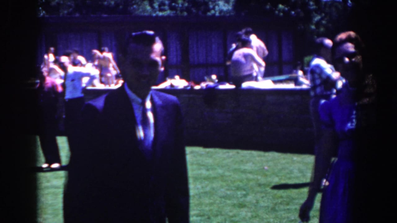 Man in a dark suit walks on a lawn beside a woman in a blue dress at an outdoor party.