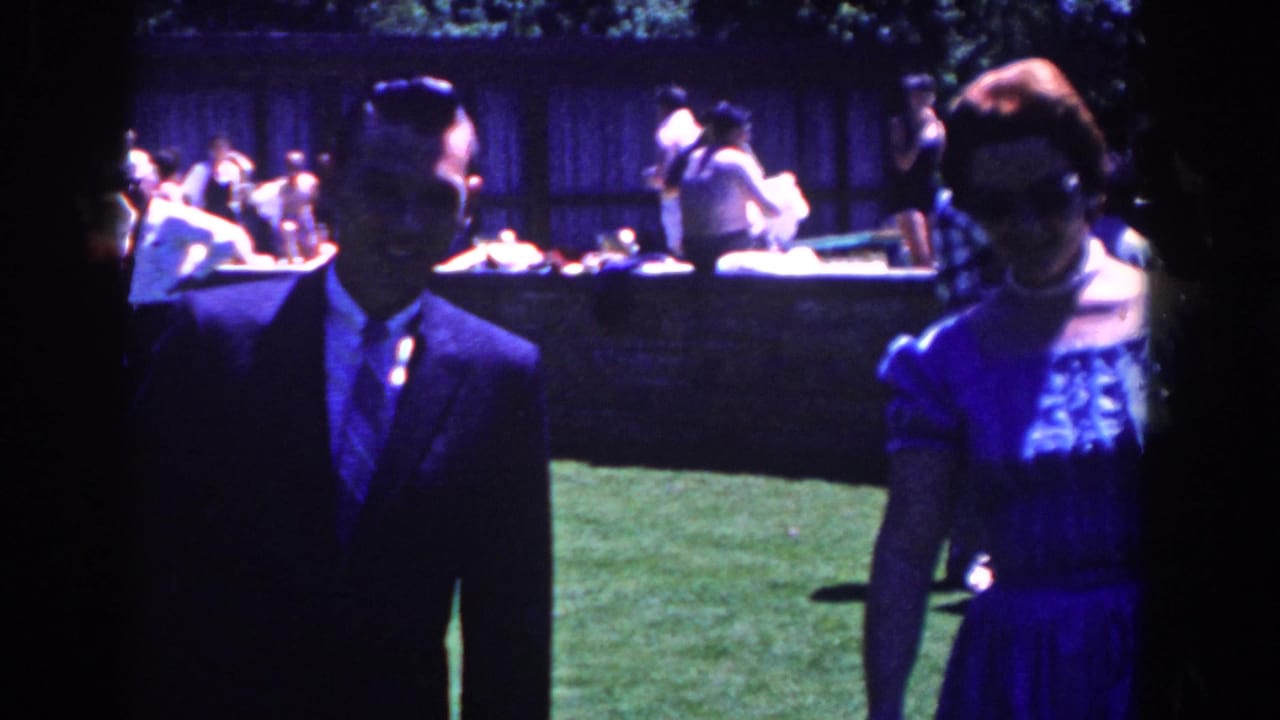 A suited man and woman in a blue dress stand near the camera with party guests behind them.
