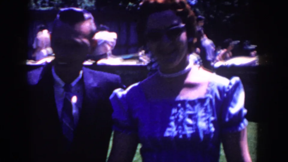 The couple faces the camera in soft, shadowed 8mm footage, with a lawn party and long table visible behind them in French Lick.
