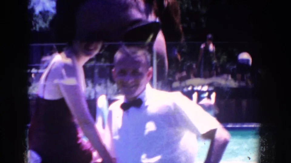 A young boy in formal clothes stands near an adult at the edge of the outdoor gathering, with water and guests visible behind him.