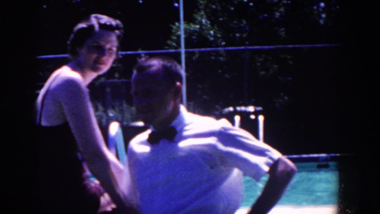 Woman in a dark bathing suit sits beside a man in a bow tie near a pool at night.