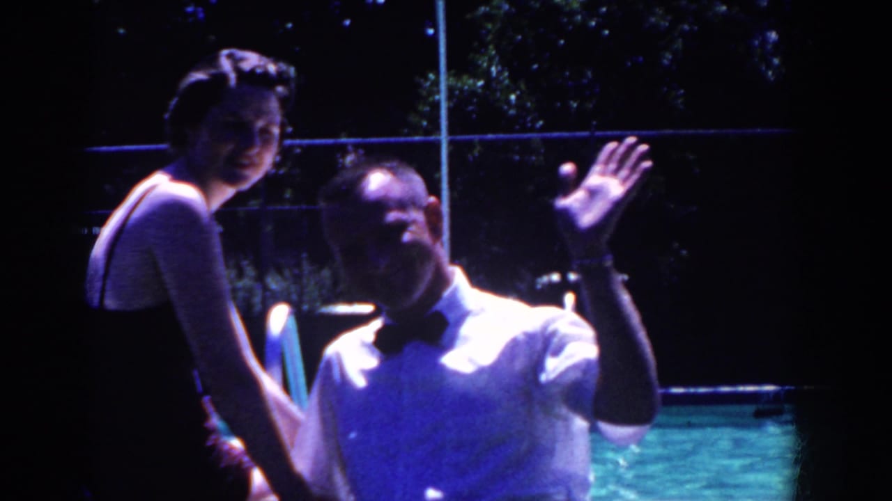 Man in a white shirt and bow tie waves beside a seated woman near a pool.