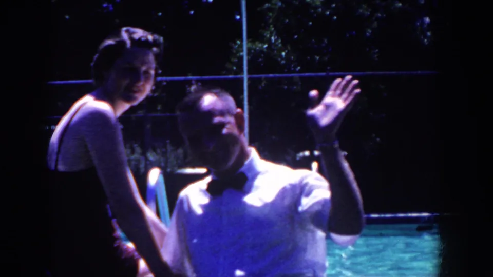 In the 1958 French Lick home movie, the man raises a hand toward the camera while the woman sits smiling beside the pool.