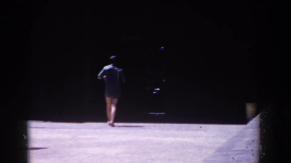 In 1958 French Lick footage, a bather in dark swimwear walks away from the camera across a sunlit beach or paved area.