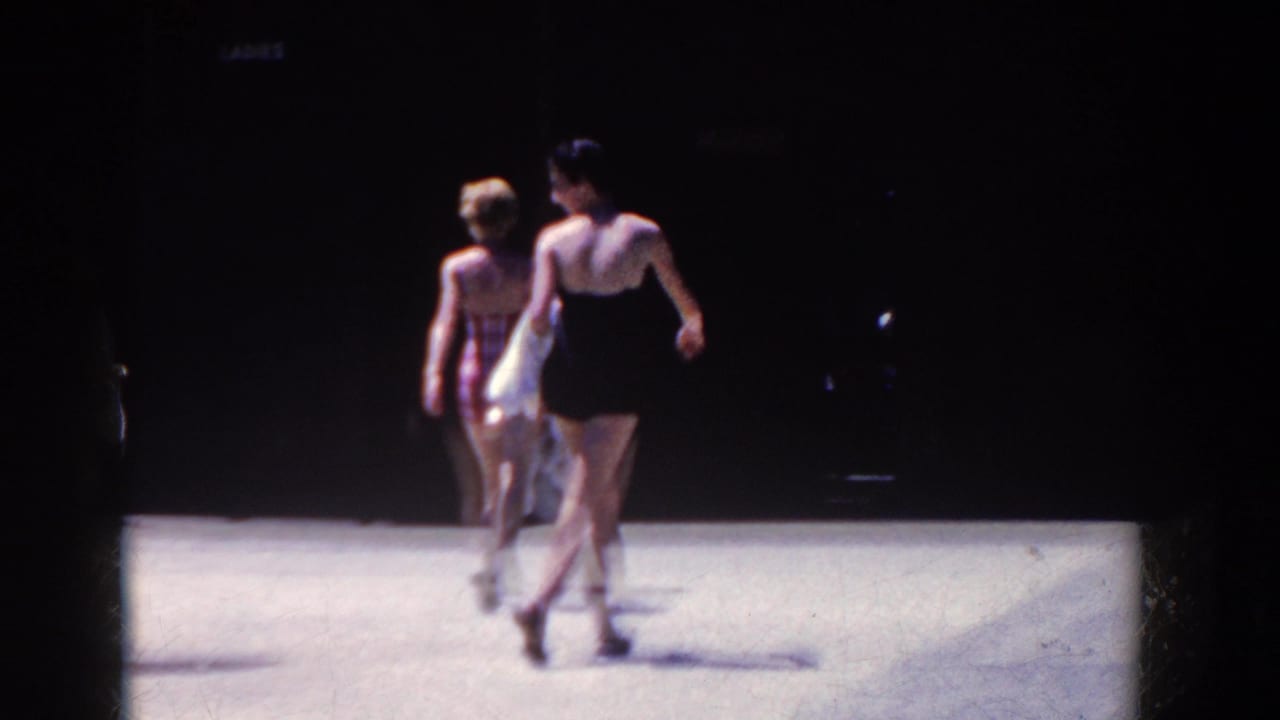 Two women in swimsuits walk away across a bright surface, one in black and one in light-colored swimwear.