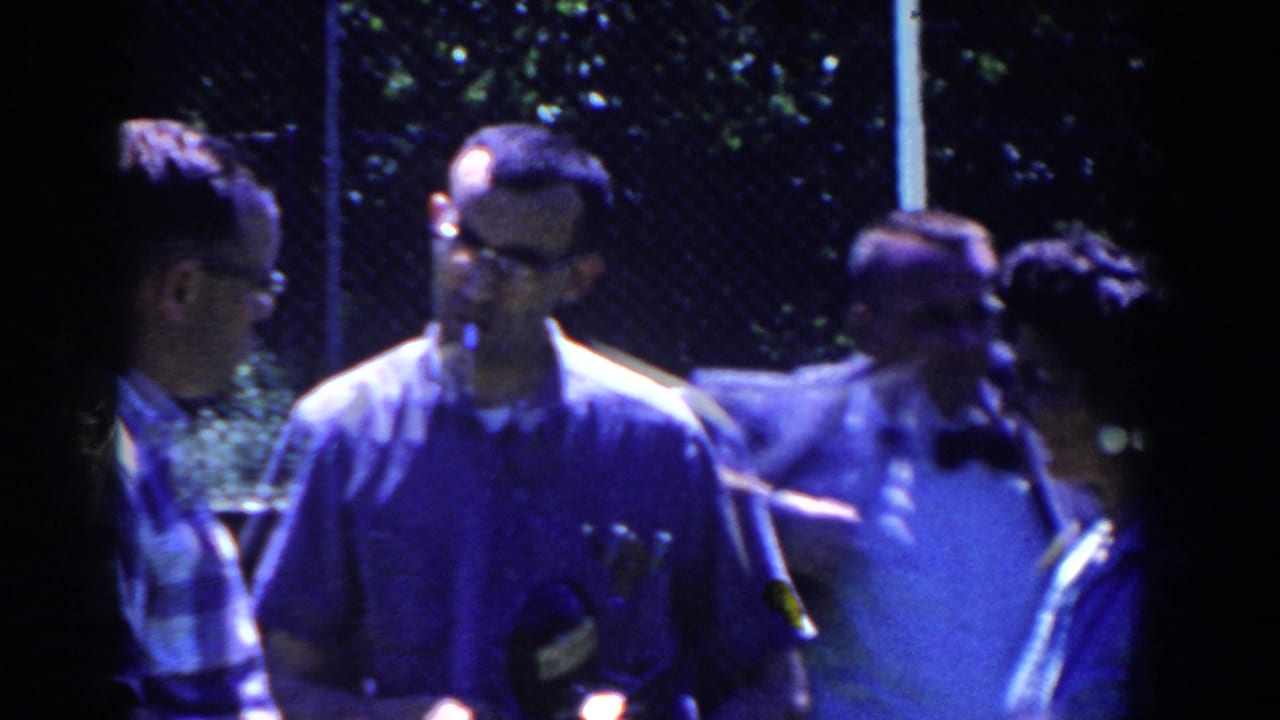 Men in casual shirts stand near a chain-link fence, with one man smoking.