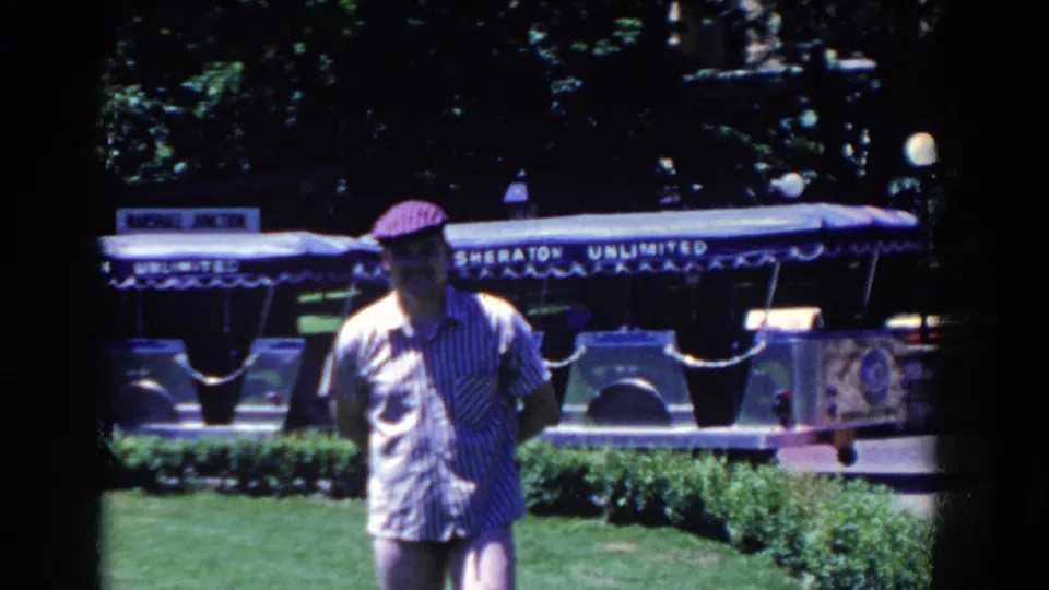 The same capped man stands in front of the Sheraton Unlimited tram, with lawn, shrubs, and trees around the outdoor area.