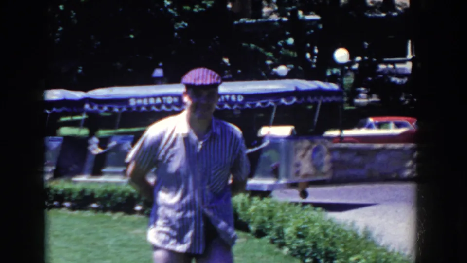 A capped man starts walking across the lawn, with the Sheraton Unlimited tram, greenery, and a small red-and-white object visible behind him.