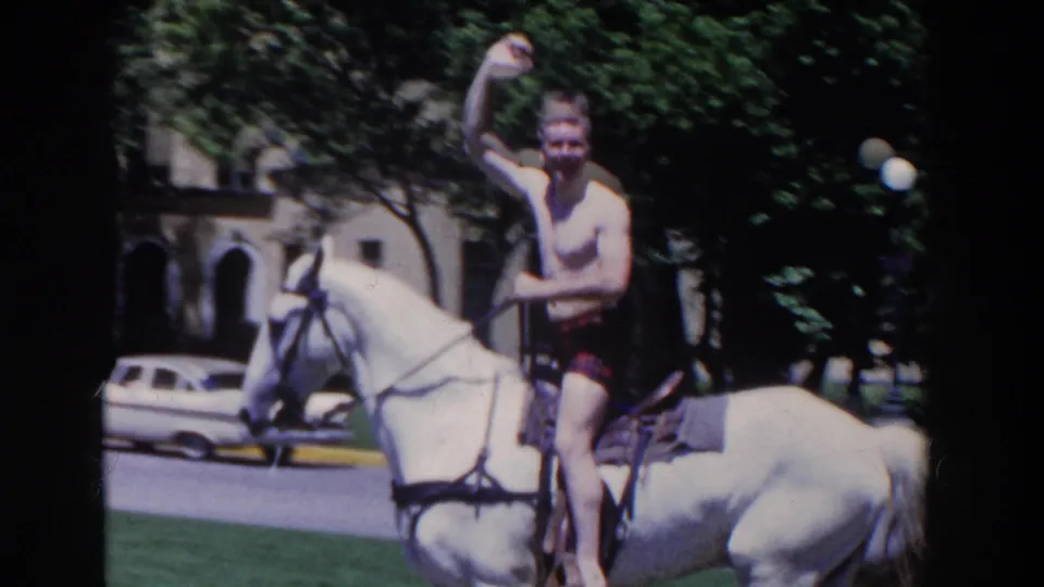 A shirtless rider raises his hand from a white horse, with a building and parked car behind him in 1958 French Lick.