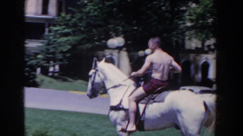 The rider is seen from behind on a white horse, moving past grass, trees, lampposts, and a background building in French Lick.
