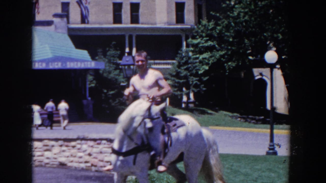 Man on a white horse faces the camera near a hotel awning and lawn.