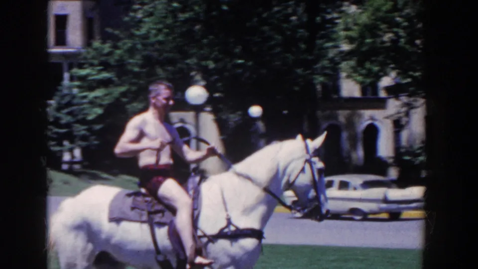 A shirtless man in red shorts rides a saddled white horse across the French Lick hotel grounds, with trees, lamps, and a mid-century car behind.