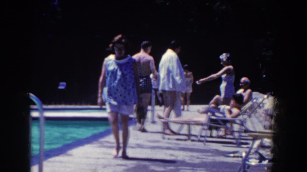 Woman in a blue patterned blouse walks barefoot beside an outdoor pool as guests sit and stand nearby.