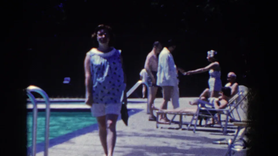 The woman in a blue patterned blouse nears the camera on a French Lick pool deck, with mid-century swimmers and lounge chairs behind her.